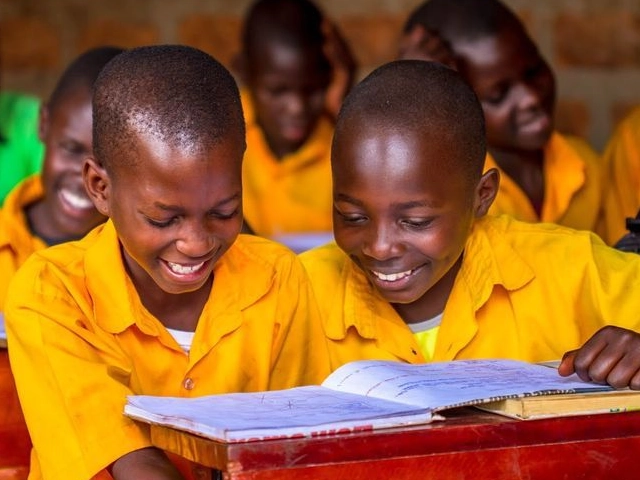 two young boys sitting at a desk reading a book and smiling